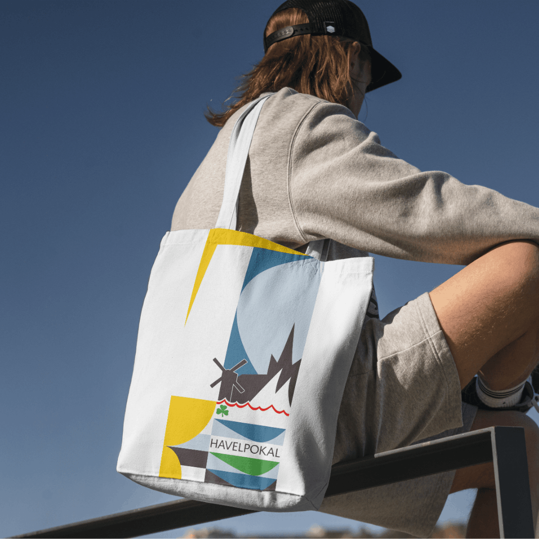 Person in Freizeitkleidung sitzt seitlich auf einer Metallstange, trägt weiße Shopper Bag locker über der Schulter. Fokus ist auf der Tasche. Die zeigt ein grafisches Design mit abstrakten, farbigen Flächen in Gelb, Blau und Braun sowie stilisierten Elementen wie Wellen, Segeln und der Stadt Werder (Havel). Zentral ist der Schriftzug 'HAVELPOKAL' platziert. Das Designsystem wirkt modern, sportlich. Es wurde von Silja Wolff entwickelt für einen crossmedialen Kampagnen Pitch.